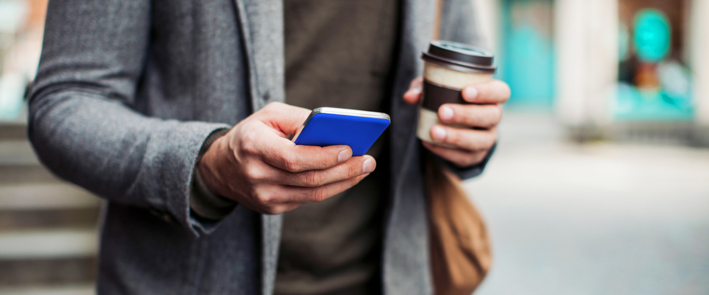 Mann mit Jacket, einem Handy in der einen und einem Kaffeebecher in der anderen Hand.