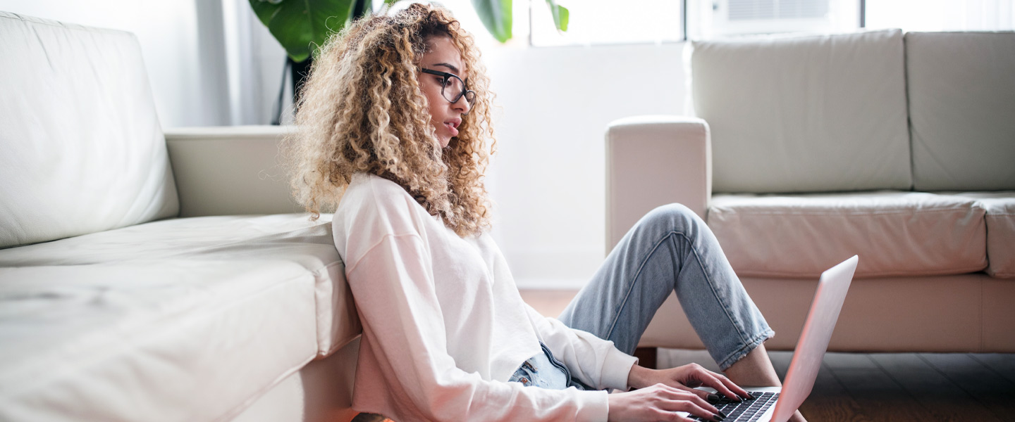 Eine Frau sitzt vor ihrer Couch und tippt auf ihrem Laptop.