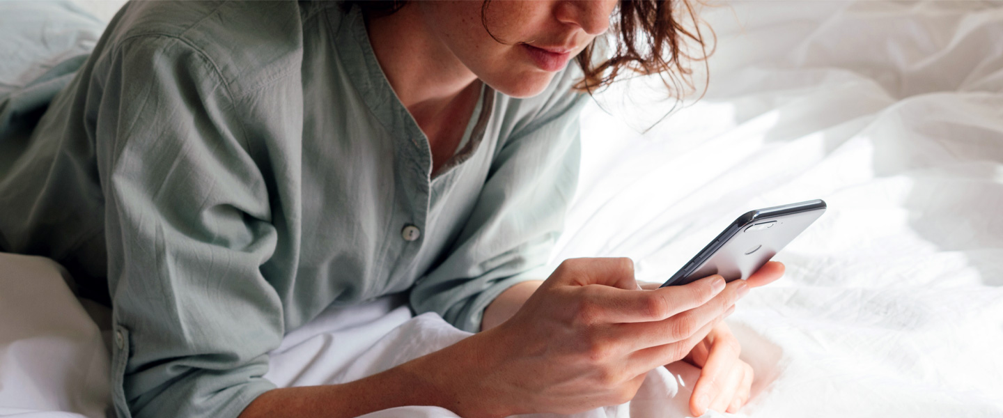 Eine brünette Frau liegt auf dem Bett und schaut auf ihr Smartphone.